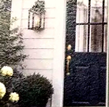 A white clapboard entrance with a navy panelled door, a hanging lantern, boxwood and white hydrangea.